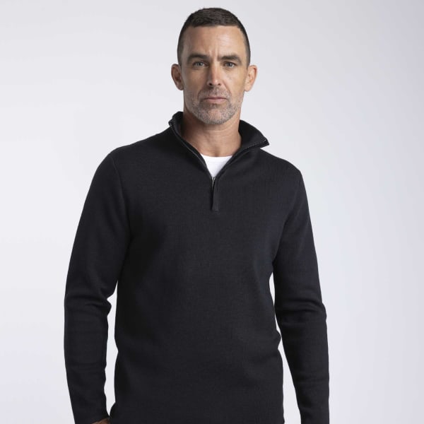 A men’s black Merino shirt with a zip collar, featuring a fitted design and long sleeves. The model stands against a plain background.