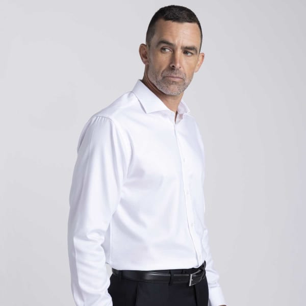 A men's button-up shirt in white, featuring a collar and long sleeves, worn with black trousers. Includes a logo.