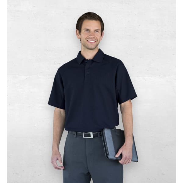 A man wearing a navy polo shirt stands against a light grey background, holding a tablet. The shirt features a logo.