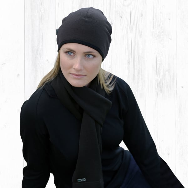 A woman wearing a black Merino Arctic Scarf and matching beanie, seated against a light wooden background.