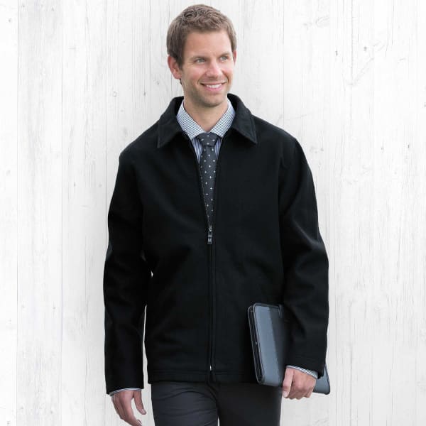 A man wearing a black Melton wool jacket, standing against a light wooden background, holding a tablet.