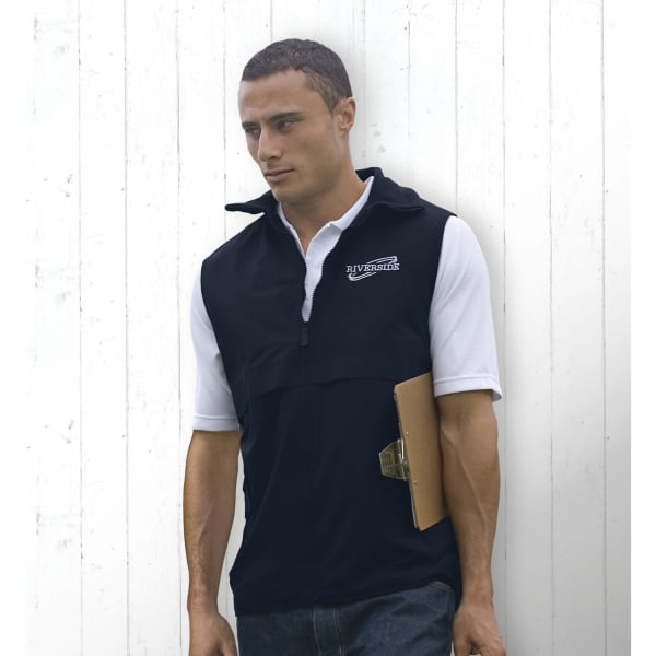 A man in a black nylon vest with a logo, holding a clipboard against a white wooden background.