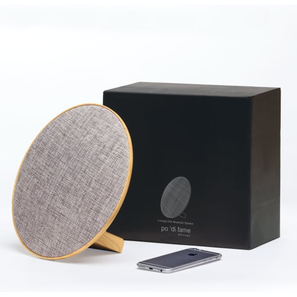 A round Bluetooth speaker with a textured grey surface and natural wood base, beside a smartphone and its black box.
