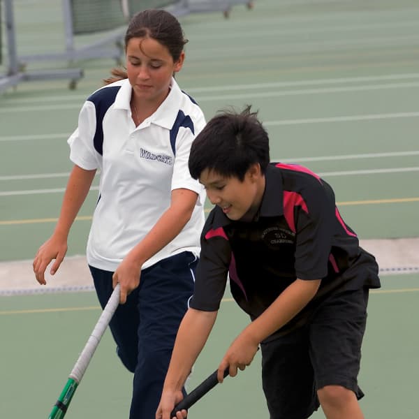 A white and black youth polo shirt with a logo, worn by a girl and a boy on a tennis court.