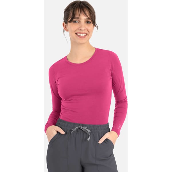 A woman wearing a long-sleeve, bright pink underscrub tee and grey pants, smiling while posing with her hands in her pockets.
