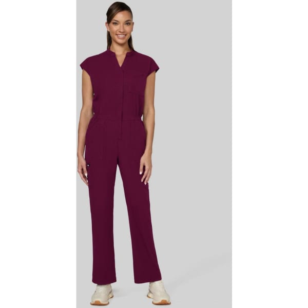 A woman stands in a burgundy jumpsuit with cap sleeves, featuring a front zip and pocket details.