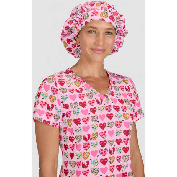 A pink bouffant cap with a heart print, worn by a model in a matching scrubs top with a heart design.