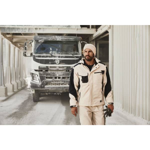 A man wearing a white and black Craftsmen softshell jacket stands in a construction setting, with a truck in the background.