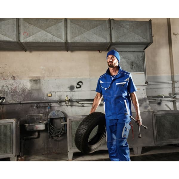 A worker in a blue workwear vest holds a tyre and a wrench in an industrial setting. The background features machinery and tools.