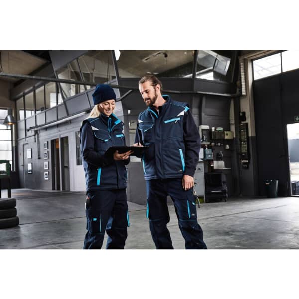A padded work vest in navy blue with teal accents, worn by two people in a workshop setting, one with a clipboard.