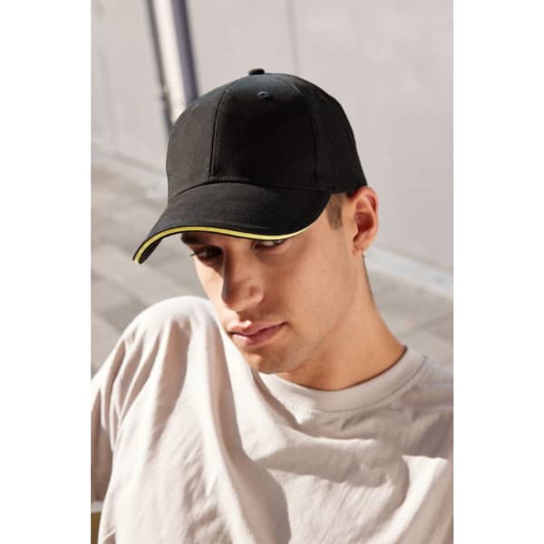 A black 6 panel sandwich cap with a yellow trim, worn by a young man in casual attire.