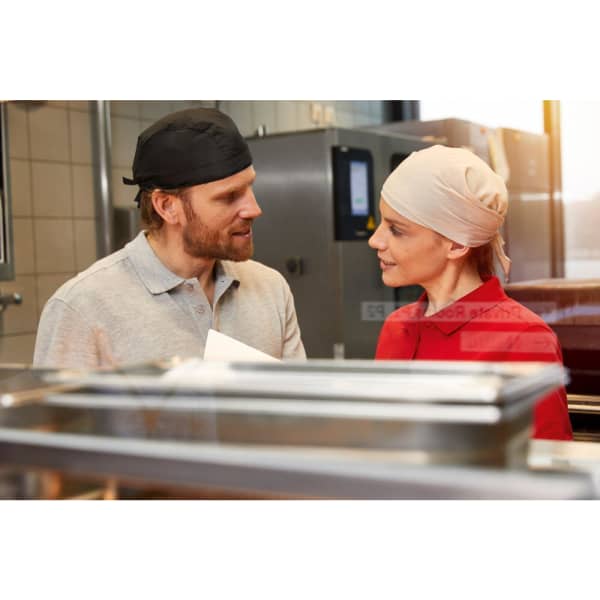 Two individuals in a kitchen setting wearing headwear, engaged in conversation, with food preparation equipment visible in the foreground.