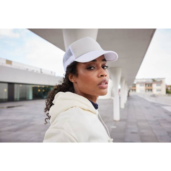 A woman wearing a white 5 Panel Polyester Mesh Trucker cap, with a light-coloured hoodie, standing against a modern backdrop.
