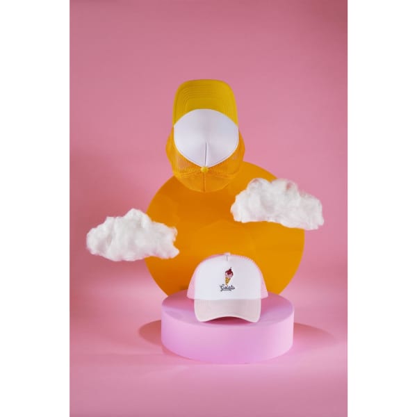 A white kids' trucker cap with a logo, displayed with yellow and pink geometric shapes and fluffy clouds.