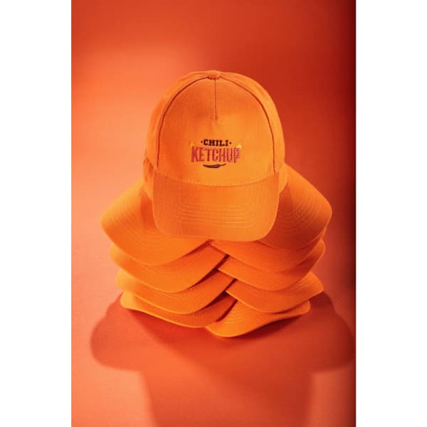 An orange 5-panel cap is displayed on top of a stack of similar caps, featuring a logo on the front.