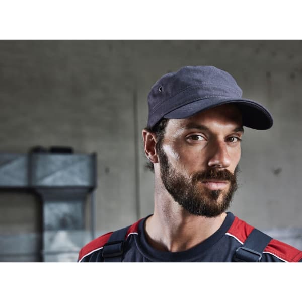 A military cap in dark grey worn by a man with a beard, set against an industrial background.