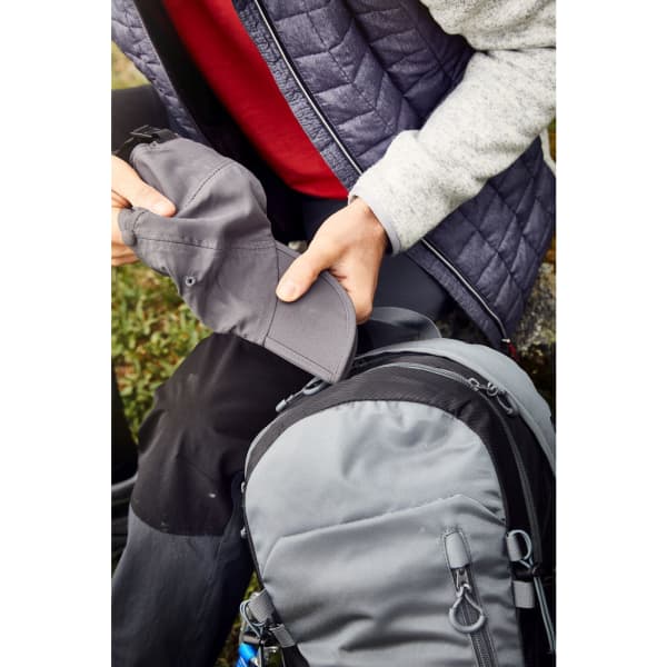 A person holds a grey cap while seated next to a grey backpack, set against a natural background.