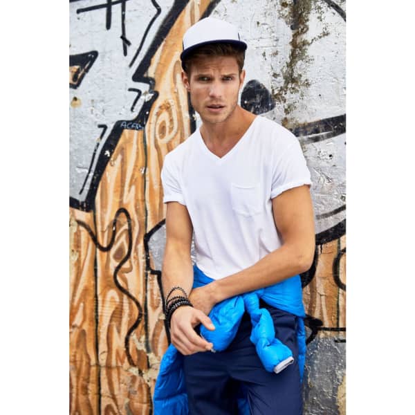 A man wearing a 5 panel flat peak cap in white, paired with a white t-shirt and a blue jacket, poses against a graffiti backdrop.
