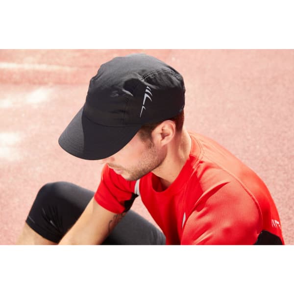 A black 3 Panel Cap is worn by a man in a red shirt, sitting on a track surface. The cap features a logo.
