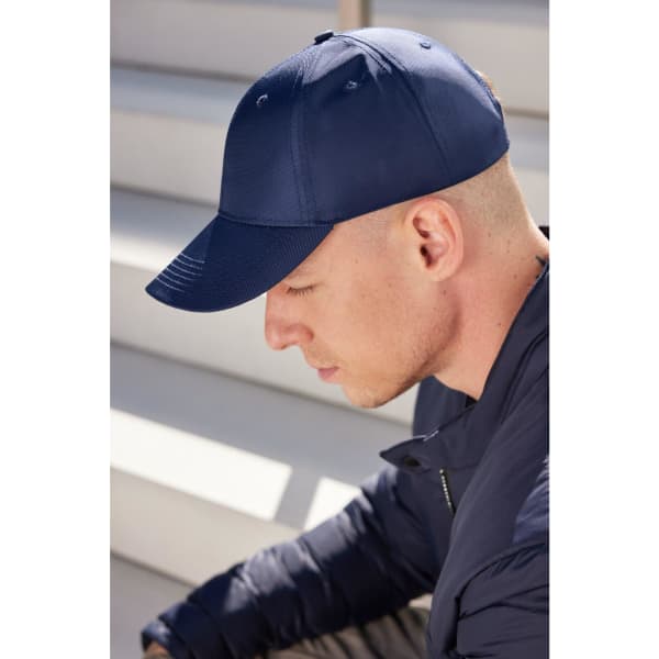 A navy blue 6 panel cap is worn by a man with a shaved head, sitting on stairs. The cap features a logo.