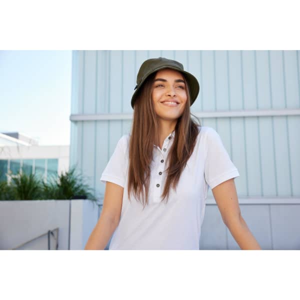 A smiling woman wearing an olive green fisherman hat and a white polo shirt outdoors, with a modern building in the background.