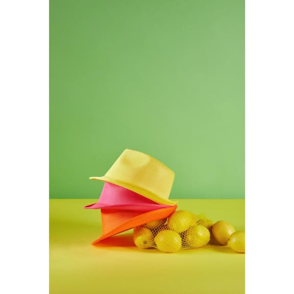 A yellow, pink, and orange hat stacked on a yellow surface, with lemons beside them against a green background.