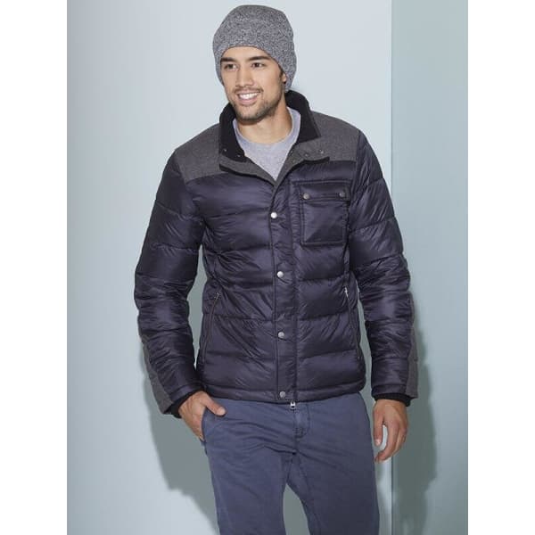 A man wearing a grey Melange beanie and a black puffer jacket, posing against a light blue wall.