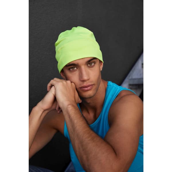 A running beanie in neon yellow worn by a model in a blue tank top, seated against a dark background.