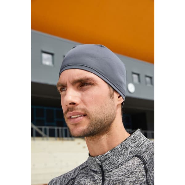 A grey running beanie worn by a man, set against an orange background with a modern building.