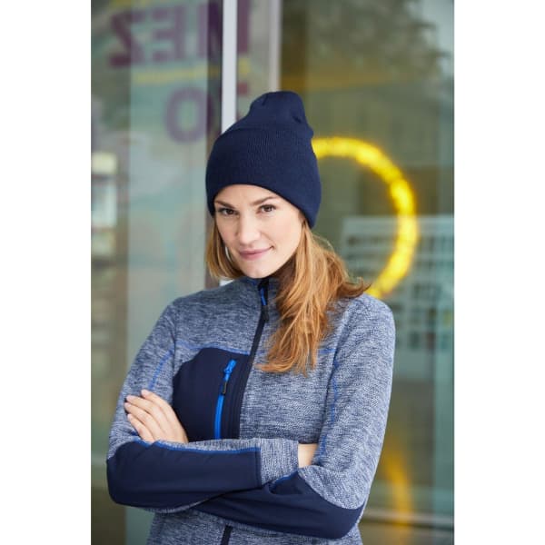 A woman wearing a navy high brim beanie and a grey zip-up jacket stands with her arms crossed. The background features a blurred circle of light.