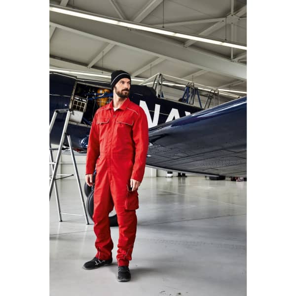 A man in a red jumpsuit and black beanie stands next to an airplane in a hangar. The image features a logo on the beanie.