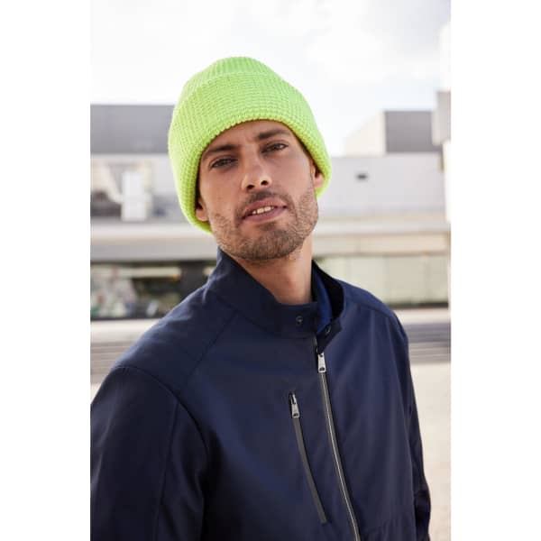 A bright yellow reflective winter beanie worn by a man in a navy jacket, set against an urban backdrop.