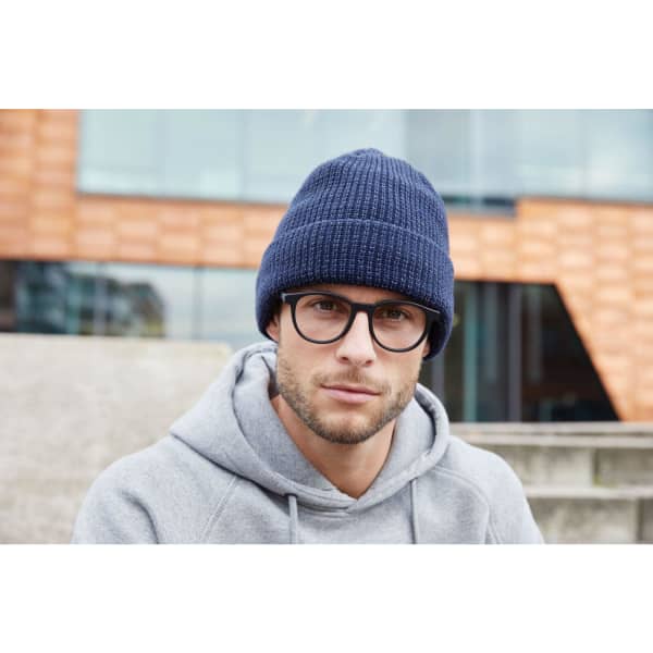 A reflective winter beanie in navy blue, worn by a man in glasses and a grey hoodie, sitting outdoors.
