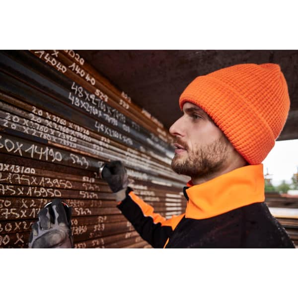 A man wearing an orange reflective winter beanie stands near metal pipes, taking notes in a construction setting.