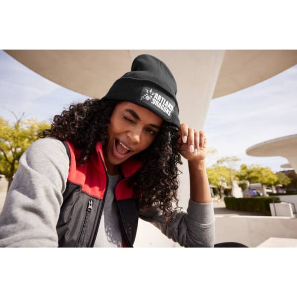 A black knitted beanie with a patch is worn by a person with curly hair, smiling and winking outdoors.