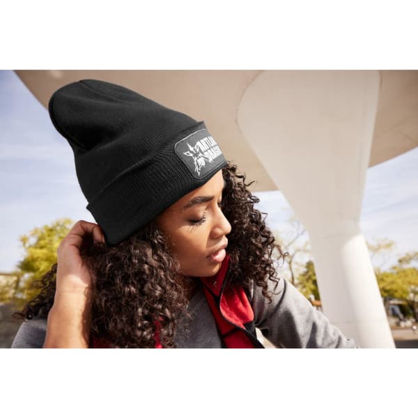 A black knitted beanie with a patch, worn by a woman with curly hair, under a modern architectural structure.