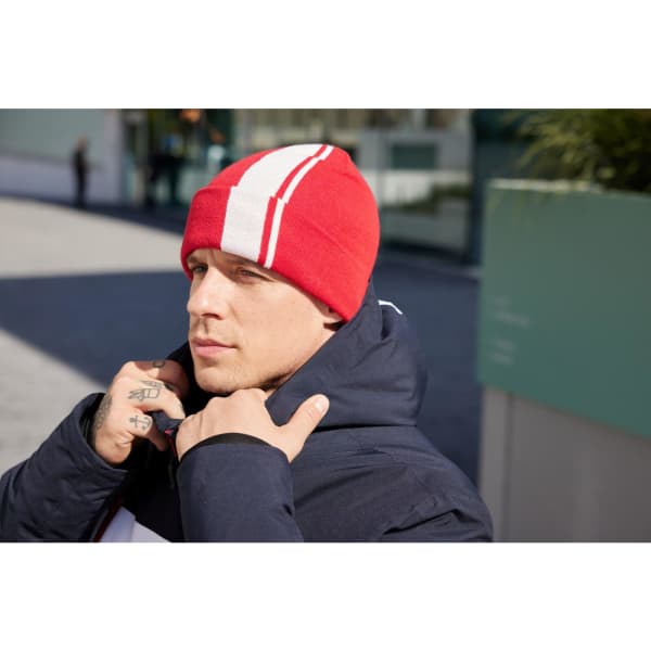 A red winter beanie with white stripes, worn by a person in a dark jacket, set against an outdoor background.
