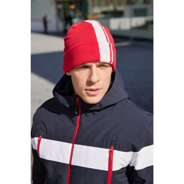 A red winter beanie with white stripes, worn by a person in a navy jacket. The background shows an outdoor setting.