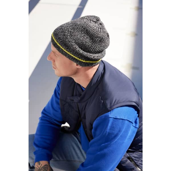 A coarse knitted beanie in grey with a yellow band, worn by a person in a blue sweater and a navy vest.