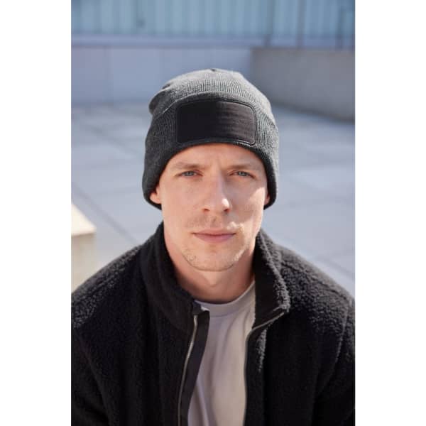 A man wearing a black beanie with a patch, paired with a dark fleece jacket, sits outdoors.