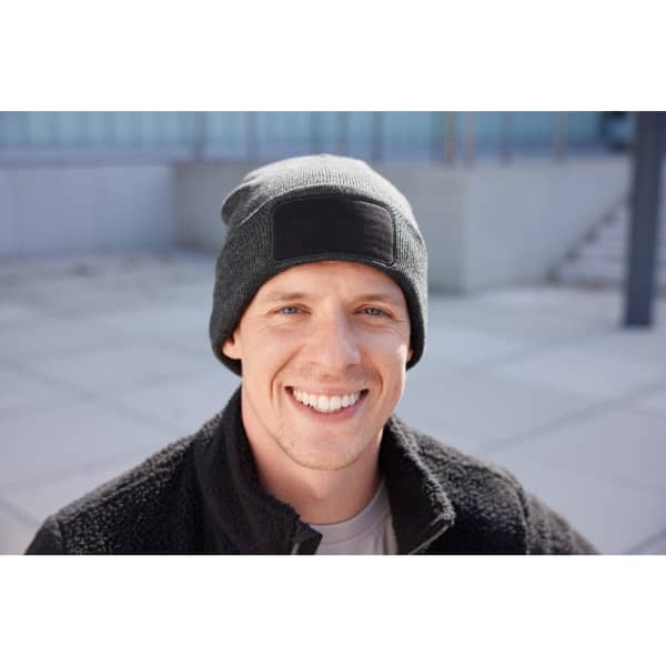 A black beanie with a patch, worn by a smiling man against a modern outdoor backdrop.
