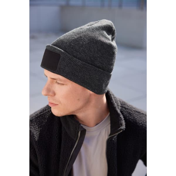 A beanie with a black patch, featuring a dark grey knit fabric, worn by a model.