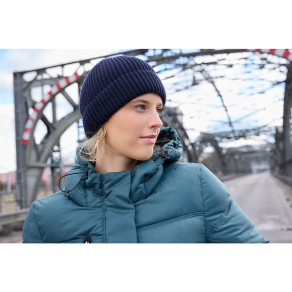 A navy blue knitted winter beanie worn by a person in a teal puffer jacket, set against an industrial bridge backdrop.