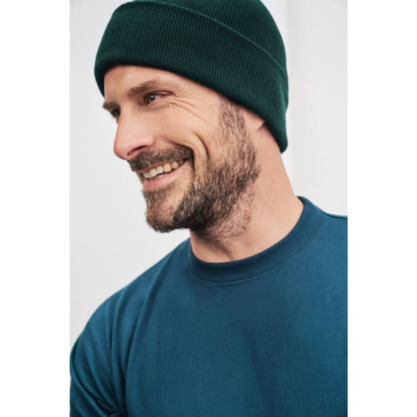 A knitted beanie in dark green worn by a smiling man, paired with a blue shirt. The image has a simple background.