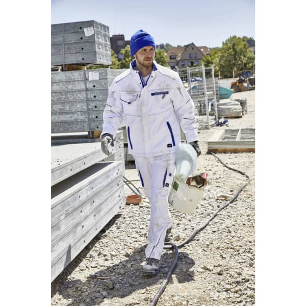 A man in a white work suit and blue knitted beanie walks on a construction site, holding a drink bottle.