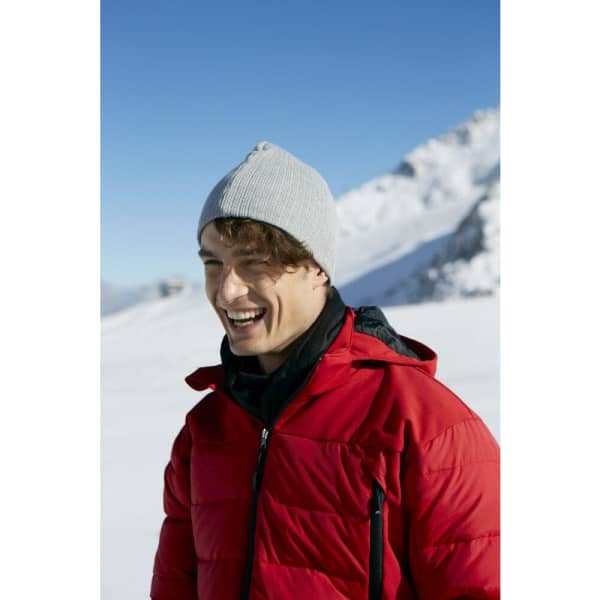 A man wearing a grey rib beanie and a red puffer jacket, smiling in a snowy mountain landscape.