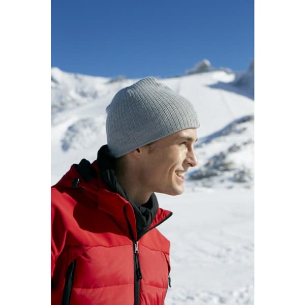 A grey rib beanie worn by a smiling person in a red jacket against a snowy mountain backdrop.