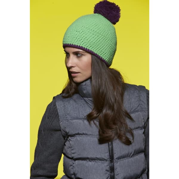 A green knitted hat with a purple pompom, worn by a woman with long hair, against a bright yellow background.