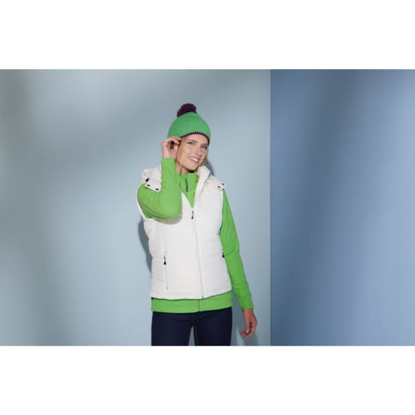 A woman wearing a white vest and a green Pompon Hat with Contrast Stripe, standing against a blue and grey background.