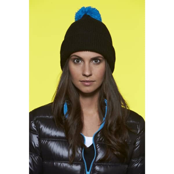 A black beanie with a blue pompom, worn by a woman in a puffer jacket against a bright yellow background.